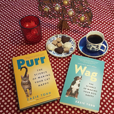 A copy of the books Purr and Wag with a plate of Christmas cookies, a red candle, a cup of coffee, and a German wooden Christmas tree with baubles on a red polka dot table cloth