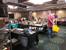 A small section of the hotel convention area with several tables, each set up with a variety of games or other products for sale. Some people can be seen either working the booth or browsing the wares.