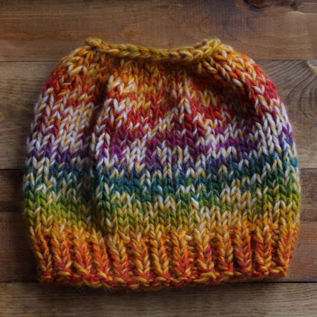 Flat shot of a yellow and rainbow knitted beanie with a hole on the top for a pony tail or bun. It lays on a wooden surface.