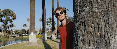 Movie still for A24's latest film Under the Silver Lake where Andrew Garfield looks sneakily from behind a palm tree with his sunglasses on