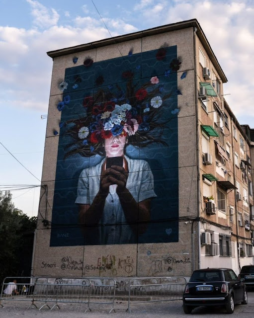 The Fabian Bane's mural in Tirana where a girl is painted looking at her phone and the upper half of her face is covered with colorful flowers