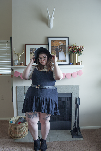 An outfit consisting of a black wide brim floppy hat, a white and black 3/4 sleeve turtleneck top under a black tiered knee length dress and black ankle boots.