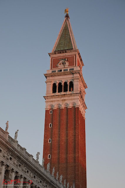 The Campanile topped with a golden angel, green tiles, white stone on red brick, the winged lion of St Mark warm and glowing in morning light, Venice, Italy.