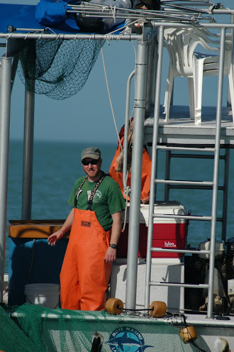 Damon is on the deck of his work boat.  There is a green net all around him and he's wearing orange Grunden overalls.