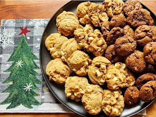 A plate full of assorted cookies on a round tray - sitting on a christmas placemat with a snowy tree on the left , on a wood table.