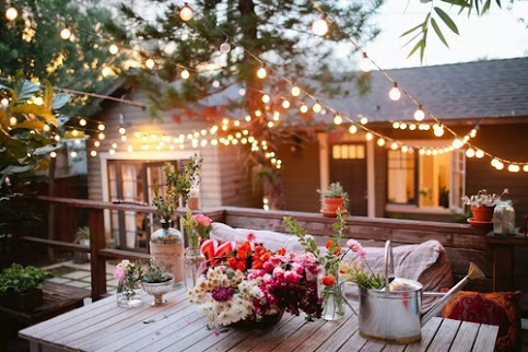 A wooden table in the foreground with flowers and a metal watering can on it . In the background are lights in the tree and a light-colored house with the windows lit up.