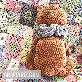 The back of a brown crochet teddy bear with a stumpy little tale, sitting on a colourful granny square blanket.