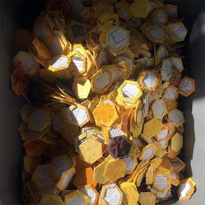 A photo from above of a grey tote box full of golden fabric hexagons basted around paper pieces, tossed like waves and brightly lit by a shaft of sunlight.