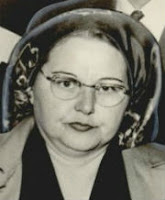 Headshot of a plump, dark-haired, middle-aged white woman wearing early 1950s style eyeglasses, hat, and hair wtyle