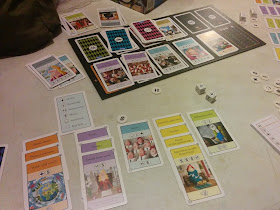 A game of CV in progress. The three decks are arranged at the top of the board, with one goal card beside them. Five cards are available to purchase along the bottom of the board. In the foreground, one player's tableau of purchased cards is visible. The special dice, can be seen, as well as the bonus tokens, scorepad, and a player aid.