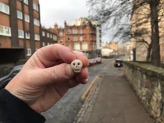 A photo of a small, ceramic skull (Skulferatu #93) being held up.  In the background is the street of Tolbooth Wynd.  Photograph by Kevin Nosferatu for the Skulferatu Project.