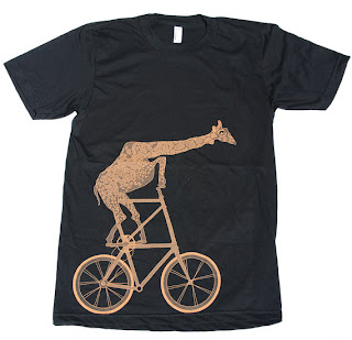 Black short-sleeve T shirt with a tan-orange print of a giraffe on a bicycle. The print takes up most of the bottom of the shirt and extends to the chest. It's comical, with a very tall seat and handle bars, and the giraffe's neck extending almost horizontally across the shirt.