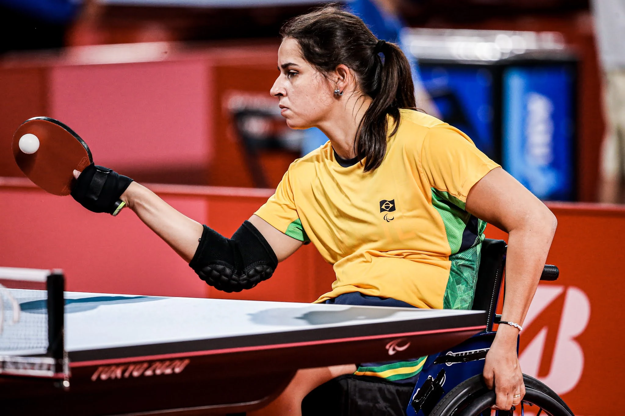 A mesa tenista Cátia Oliveira, de amarelo, rebate a bola branca com a raquete vermelha na mão direita, enquanto a outra mão está segurando a roda da cadeira de rodas, na qual a atleta está sentada