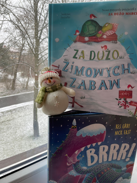 Na okiennym parapecie stoją pionowo, jedna na drugiej, dwie książki o zimie: "Za dużo zimowych zabaw", "Brrr: gdzie zniknęły dinozaury?". Na jednej z książek siedzi pluszowy bałwan w czapce i szaliku. Za oknem widać sypiący śnieg.
