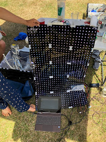 fifteen LED grids taped into a rectangle, held up by someone wearing pyjama pants, in front of a cluttered table, resting on grass