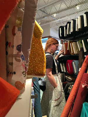 A shot down the crowded aisle of a fabric store, slightly obscured by fabric draping off bolts on the left, showing a young white woman with long, braided red hair, inspecting stacked bolts of thick fabric. She's wearing a black t-shirt and white suspender skirt with a faint blue-outlined shell pattern and large pockets topstitched in red.