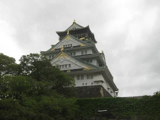 Osaka castle. Photo taken from below. A five story white walled building with green colored roof on each floor with gold statue at the front.