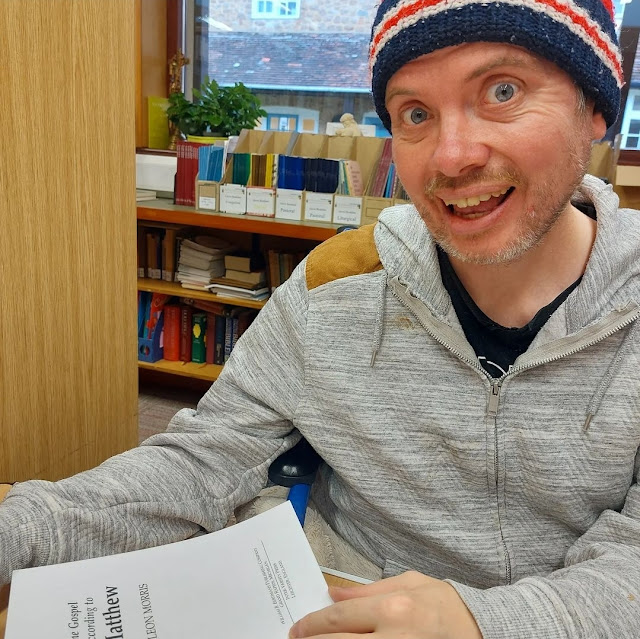 Martyn wears his red and black woolly hat, is beaming at the camera while srudying a college libray room. He is wearing a grey hoody and black tshirt