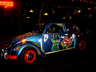 This is a picture of a classic blue beetle painted with images.  This is a side view the images that can be seen are a green nude lady and red mushroom behind the car can be seen the lights of the Whiskey Girl Tavern.