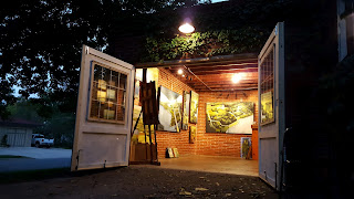 An image of Danielle Clouse Gast's home gallery space, with the doors wide open and bright lights on inside the building. Artwork is hanging on the walls inside.