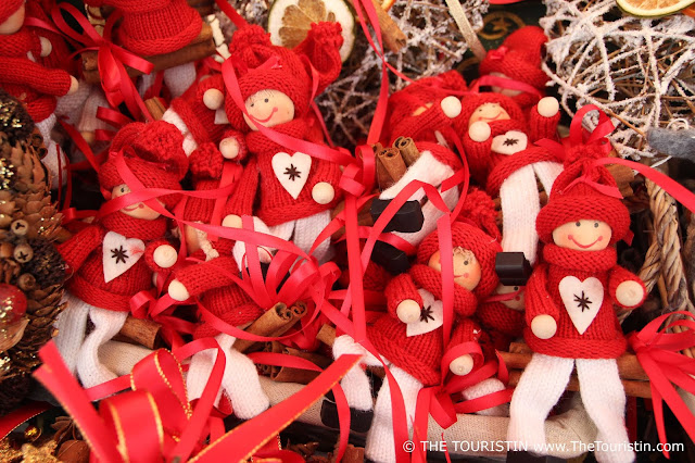 A bowl with a group of small brightly smiling knitted figurines wearing a red beanie, black wooden booties, white trousers, and red jumpers with a white felt heart on its front.