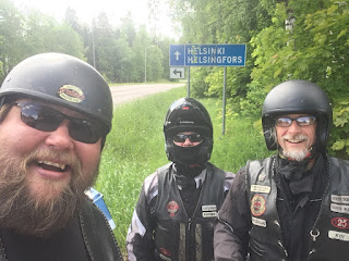 Three bikers taking a selfie in front of a signpost for Helsinki.