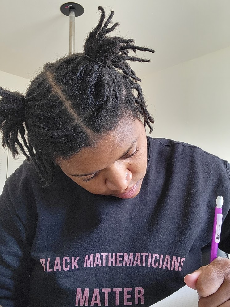 Black woman with locs in 2 ponytails wearing a crewneck that says "Black Mathematicians Matter" She has her head down and is holding a purple mechanical pencil in her left hand.
