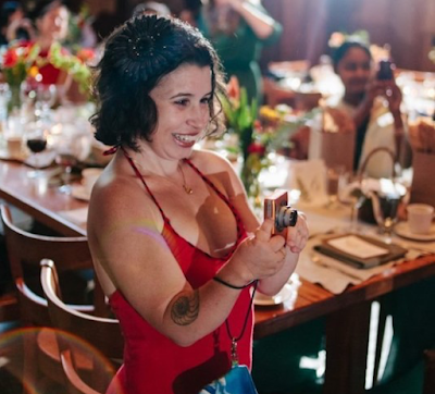 Small Latinx white woman in beight red dress, short dark hair, smiling, and taking photos of a crowd at a party.