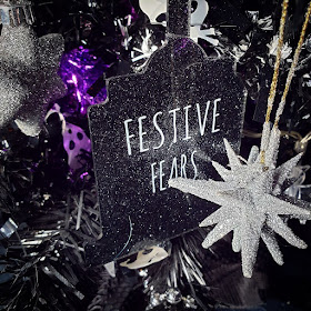 A black gilltery tombstone-shape cut from acrylic, with a transfer in white stylised spindly letters saying 'Festive Fears', suspended in a black Christmas tree, with a white star bauble in the foreground, and a grey six-pointed star bauble to the left. There is skull tinsel in the background