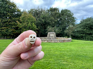 A photo of a small ceramic skull (Skulferatu #109) being held up.  In the background is the stone memorial to the Battle of Roslin.  Photo by Kevin Nosferatu for the Skulferatu Project.