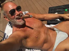 Looking down upon a sunglasses wearing grey bearded older daddy sunbathing with cigar in mouth