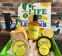 A car photo of a square light brown box containing some white ecopops, a syringe plastic shower gel bottle filled with yellow shower gel and a black with a yellow rectangular label with lemon shower gel Lush in green font next to a square slab of light yellow soap with yellow bits of lemon zest running throughout it next to a black cylindrical pot filled with slightly tinted yellow but still white body conditioner with a circular black lid and a circular yellow label with  Lemony footer body conditioner Lush in green font next to a clear plastic cylindrical tub filled with yellow and green facial cleanser with a clear circular red and a yellow circular label with lemon aid facial cancer Lush in green font next to a small cylindrical glass jar filled with yellow sugar scrub with a black circular lid and a yellow circular label with nibble lip scrub Lush in green font with a yellow and green cardboard sleeve behind the box with when life gives you lemons Lush in yellow and green font on a light brown rectangular table on a bright background