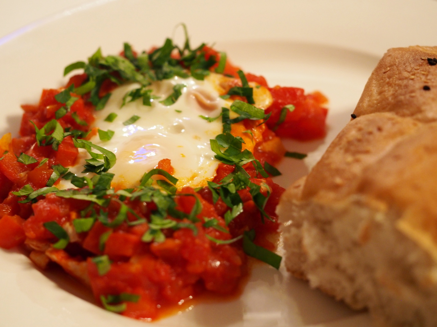 Eine Portion des Shakshuka auf einem Teller, inklusive dem poschiertem Ei in der Mitte der Tomaten-Paprika-Masse. Daneben liegt ein Stück Fladenbrot.