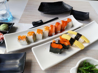 Two rectangular white plates filled with rectangular sushi with pink and red rectagnaulr strips on top on a large white table on a bright background 