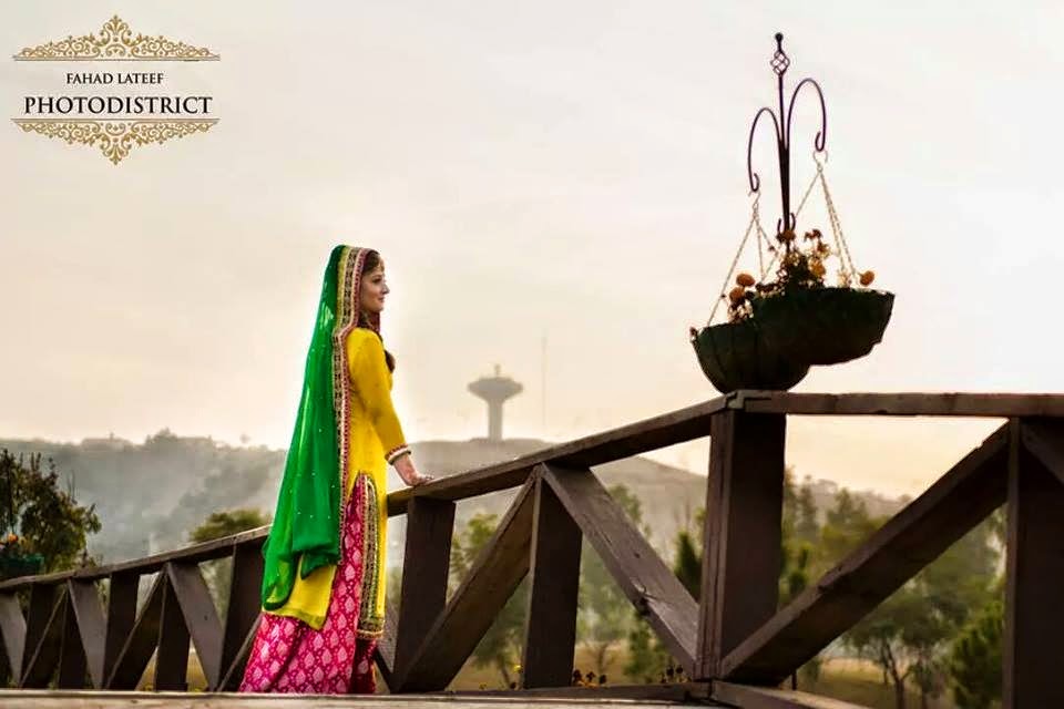 A bride standing on a deck, looking down on the grounds. Wearing a yellow shirt, a pink ghagra (traditional Skirt) with silver botis (a type of flower) embroidered on it and a green head veil