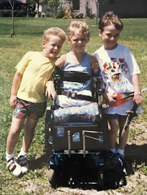 A picture of three boys, standing shoulder-to-shoulder. The middle child is a power wheelchair user
