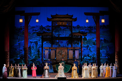 A stage with singers in sumptuous costumes lined up across it; a red gateway also stretching across the stage and very tall; a blue background displaying a village.