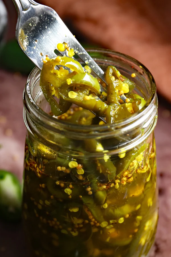 A close-up view of a jar brimming with enticing cowboy candy. A fork is delicately digging into the jar, lifting out the luscious candied jalapenos. The appealing colors and tempting textures of the jalapenos offer a delightful combination of spicy and sweet flavors, ready to satisfy any craving.