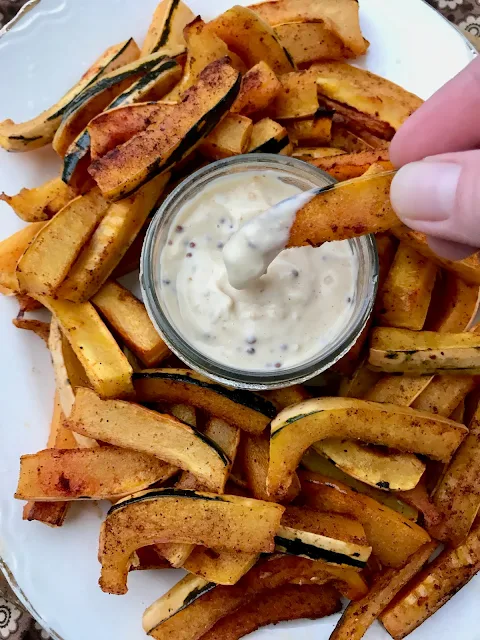 Finished roasted delicata squash sticks on a platter with dipping sauce and one stick being dipped into the sauce bowl.