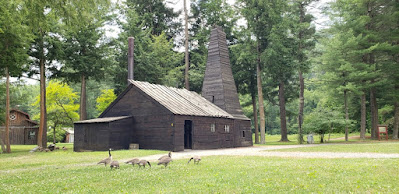 Wooden building with peaked roof and wooden tower that houses replica oil derrick. The building is set among trees and a green lawn on which 7 geese are gathered.