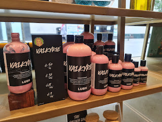 A photo showing a row of cykdienuxao clear bottoes with black circular lids filled with light pink liquid with a black label that says Valkyrie conditioner lush in white font on a light brown shelf with a black card that says Valkyrie conditioner and a range of prices in white font on a bright background