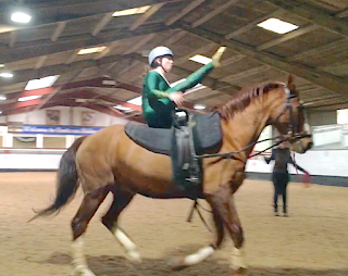 A vaulter sitting on a large chestnut gelding in canter. The horse and vaulter are side-on to the camera, and the vaulter's nearest leg (the right leg) is lifted straight in the air. The vaulter's left hand is about to regrasp the handle on the vaulting roller.