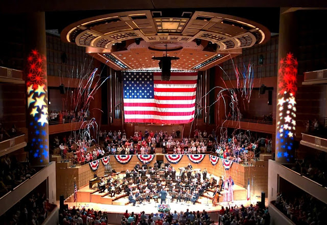 Backed by a huge US flag and surrounded by a burst of streamers and projected stars and fireworks in red white and blue, the Dallas Winds performs on the stage of the Morton H. Meyerson Symphony Center in Dallas, TX.