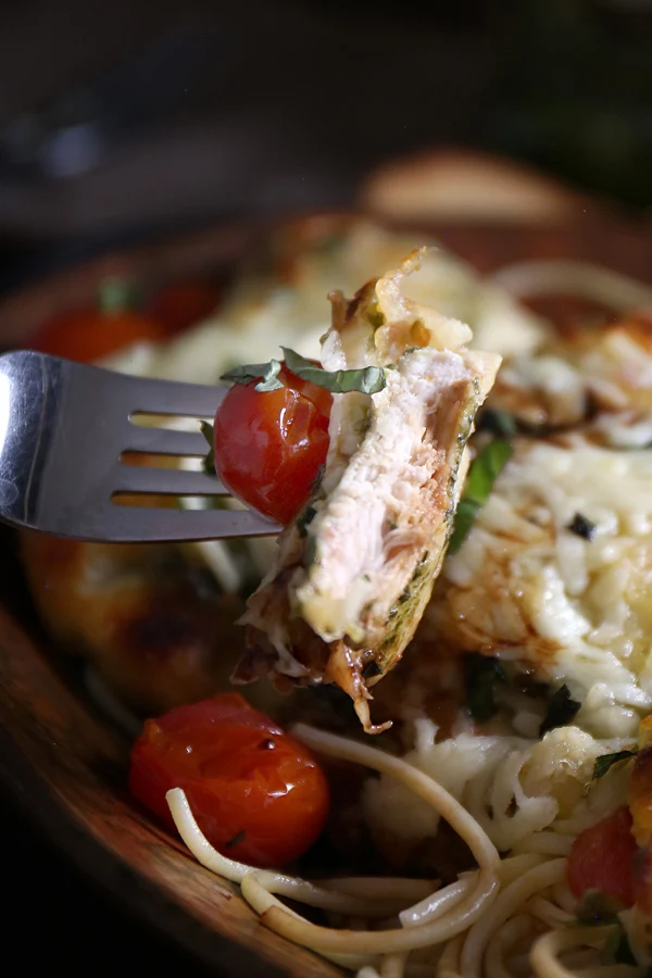 A forkful of cheesy baked pesto chicken with a roasted grape tomato and fresh basil, showcasing the juicy goodness of the flavorful chicken.