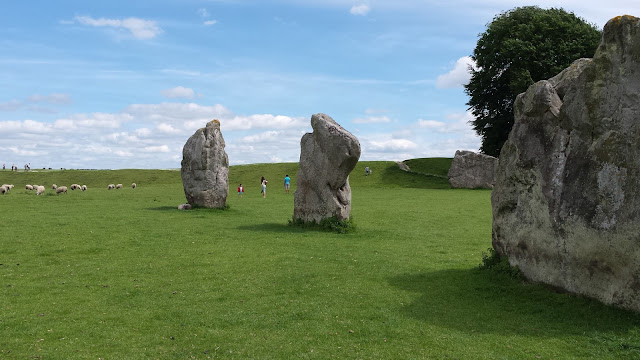 sfeerbeeld met blauwe lucht en groen gras. Hierop zijn massieve stenen te zien van de steencirkel te Avebury. Schapen en mensen lopen tussen de stenen.