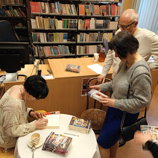 Spotkanie autorskie w bibliotece z Danutą Noszczyńską. Podpisywanie książek przez autorkę parze czytelników.