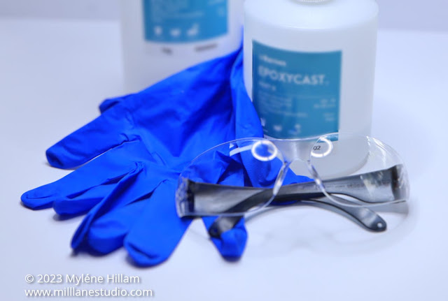 Blue nitrile gloves and wraparound safety glasses on a white table with bottles of resin and hardener.