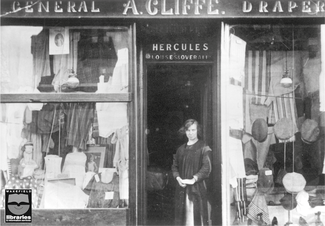 Annie Cliffe, as a young, stern-looking woman outside her Drapers Shop. The two large display windows either side of the door are filled with stock for sale. The handpainted sign above the door reads 'General - A. Cliffe - Drapers'