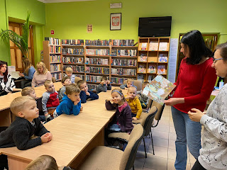 Panie bibliotekarki stoją przez przedszkolakami, jedna z nich czyta im książki.