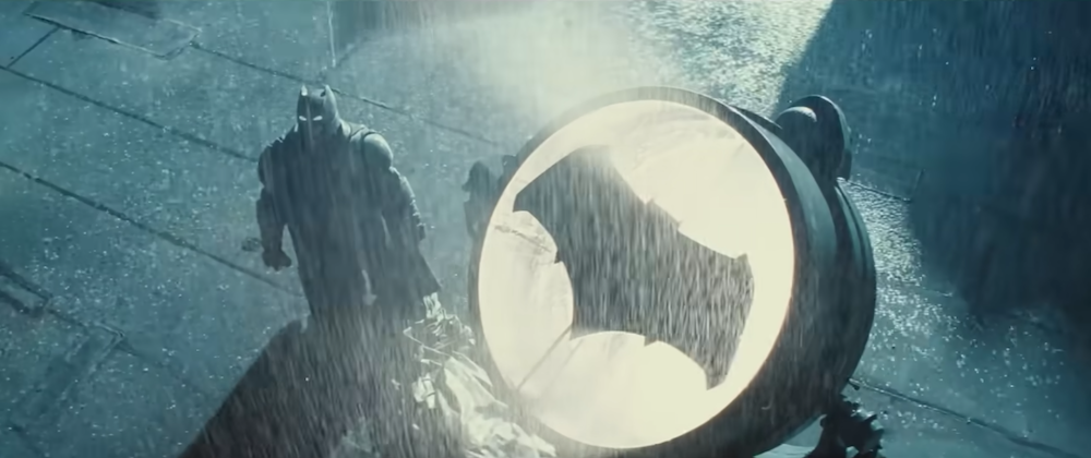 Batman standing in the rain on rooftop next to lit Bat-Signal